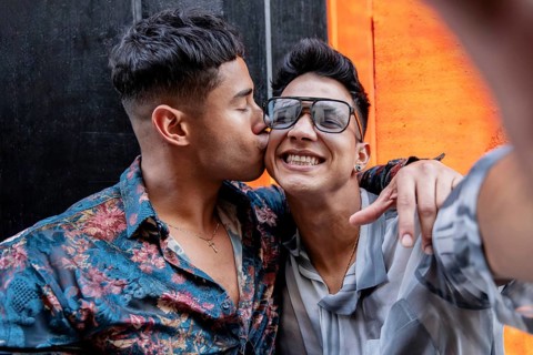Latin gay couple taking selfie while embracing warmly. One partner kisses the other's cheek, while