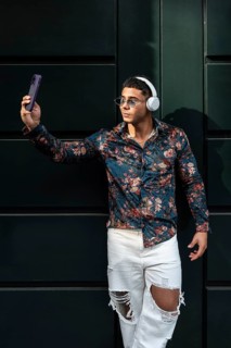 Stylish man wearing floral shirt and headphones takes a selfie. He wears sunglasses and ripped