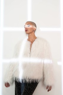 A middle-aged woman with a shaved head poses confidently, wearing a fashionable white faux fur coat