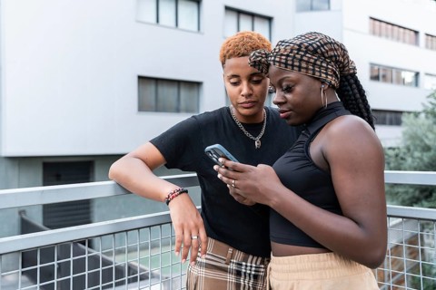 An LGBT multiethnic couple shares a tender moment outdoors, both focused on a smartphone, with one