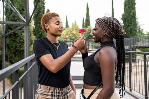 An LGBT multiethnic couple shares a tender moment in a serene park setting, exchanging a flower and