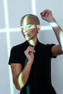 A middle-aged woman with a shaved head poses gracefully under artistic light patterns, radiating