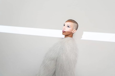 A middle-aged woman with a shaved head poses confidently, wearing a stylish furry coat against a
