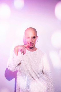 A middle-aged woman with a shaved head poses confidently, bathed in soft, bright light with lens