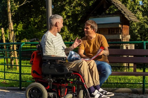 Two friends sit together on a park bench, one in a wheelchair by cerebral palsy and the other