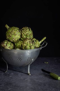 A collection of fresh artichokes sits in a rustic metal colander on a dark counter, accompanied by