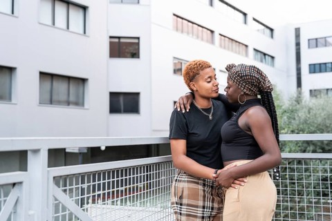 An affectionate LGBT multiethnic couple leans against a balcony railing in an urban setting,