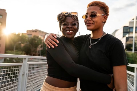 An LGBT multiethnic couple embraces warmly on a pedestrian bridge. They smile with genuine