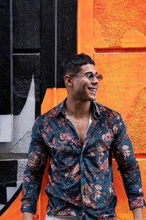 A stylish latin man in a floral shirt and sunglasses stands smiling against a vibrant orange and