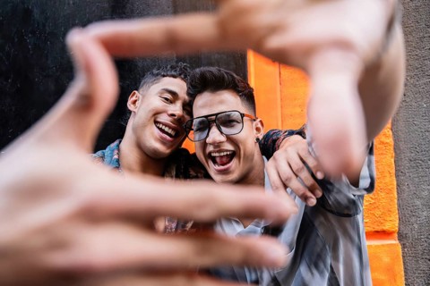 A smiling couple frames their joyful expressions with their hands against a vibrant orange urban