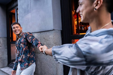 A smiling latin gay couple holds hands outside an urban cafe, radiating happiness and love. One