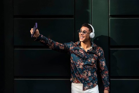 A young latin man in a floral shirt, wearing sunglasses and headphones, smiles while taking a