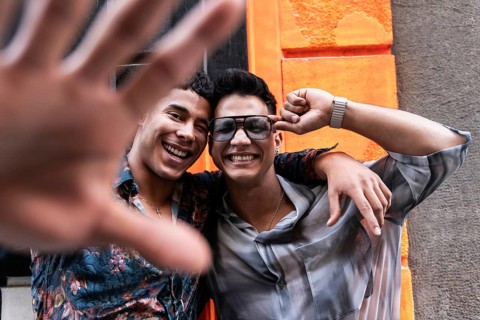 A smiling couple poses playfully in front of a bright orange wall. One partner playfully reaches