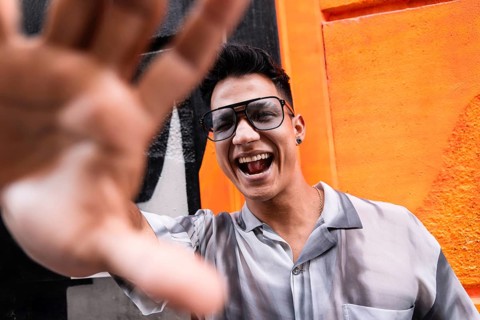 A young man wearing glasses and a stylish shirt smiles joyfully, extending his hand toward the