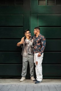 A stylish latin gay couple stands close, exchanging affectionate looks against a large dark green
