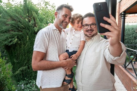 A joyful gay couple takes a smiling selfie with their son in a lush garden setting. The image