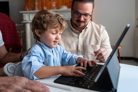 A young child types on a tablet keyboard, guided by an adult. They are sitting together at a table