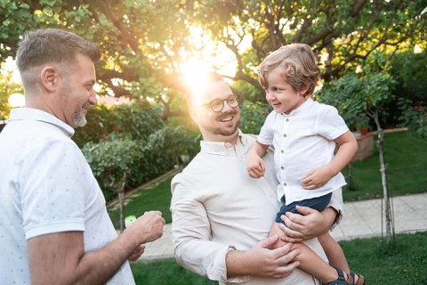 A joyful gay couple shares a tender moment with their son in a sunlit garden, surrounded by lush
