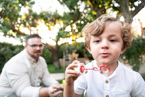 A young boy in a white shirt joyfully blows bubbles in a vibrant garden. His parent, watches in the