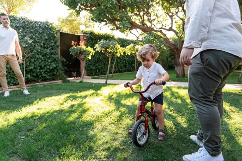 A joyful gay couple encourages their young son as he learns to ride a bicycle in a sunny garden.