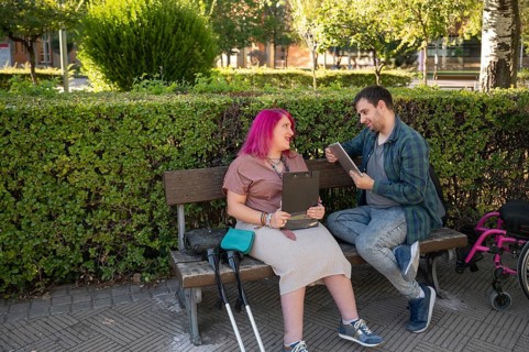 A couple sits on a park bench studying The woman, with vibrant pink hair, uses crutches due to her