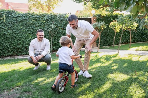 A joyful gay couple plays with their son, teaching him to ride a small bicycle in their garden. The