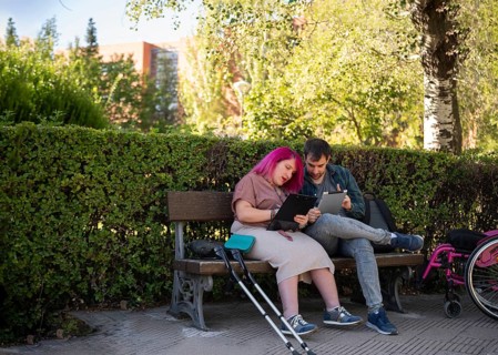 A couple studies together on a bench in a college garden The woman, with pink hair, uses a
