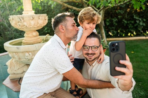 A joyful gay couple takes a selfie with their young son in a lush garden. One dad kisses the son