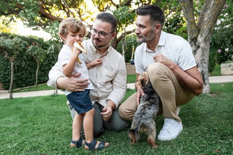 A joyful family moment in a lush garden with a gay couple watching their son eating a banana, and a