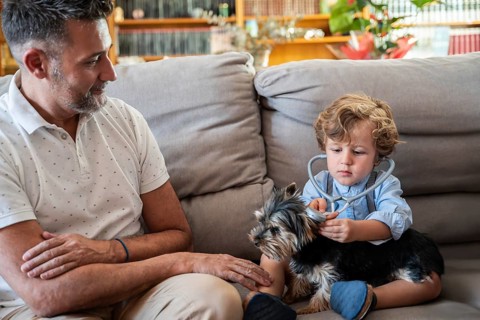 A child explores the role of a vet, using a stethoscope on a small dog, with a smiling father