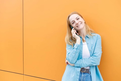 A cheerful woman stands against a bright orange wall in Warsaw, chatting on her phone She's dressed