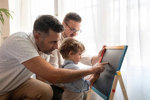 A loving gay couple engages with their young son, teaching him using a chalkboard. The warm,