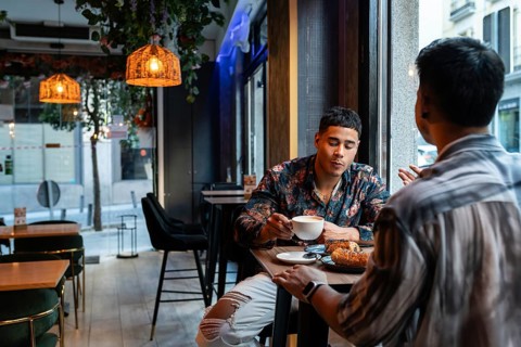 A latin gay couple enjoys a cozy moment at a café, sharing coffee and pastries. The warm lighting