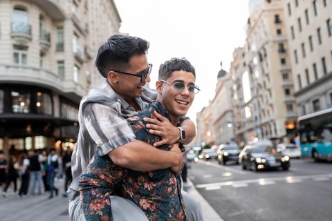 A latin gay couple shares a joyful moment on a bustling street in Madrid, surrounded by beautiful