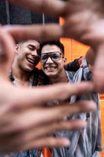 A joyful latin gay couple poses with smiles, framing their faces with their hands for a selfie