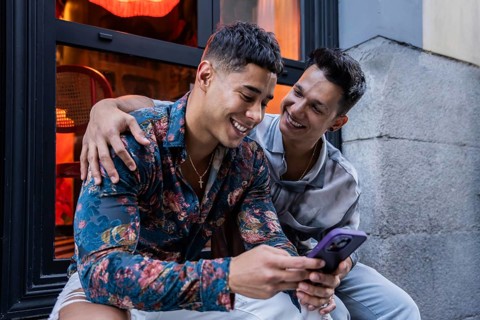 A joyful latin gay couple sits outside a cafe, sharing a moment on a phone. They are smiling and