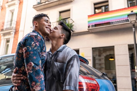 A joyful latin gay couple shares a tender moment on a lively street in Madrid, with a rainbow flag