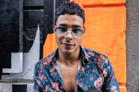 A confident latin man wearing sunglasses and a floral shirt poses against a vibrant, colorful