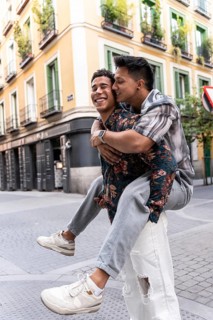 A joyful latin gay couple piggybacking on a picturesque street in Madrid. The colorful buildings