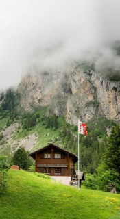 A cozy wooden chalet stands amidst lush greenery, with misty cliffs in the background at Lac de
