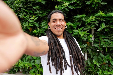 A cheerful man with long dreadlocks smiles while taking a selfie in a lush green garden in