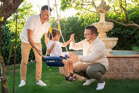 A joyful gay couple engages with their young son in a sunny garden, sharing laughter and high-fives