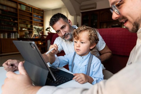 A gay couple sits with their young son in a cozy living room, focused on a tablet. They are sharing