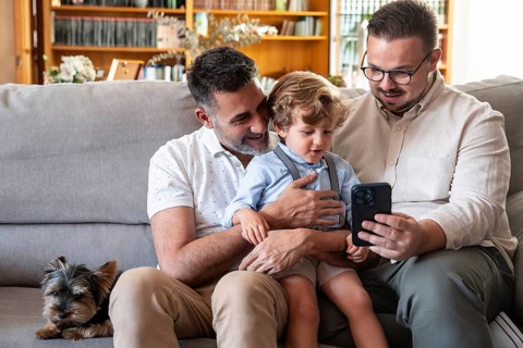 A joyful gay couple sits on a cozy couch with their young son and a small dog, sharing moments