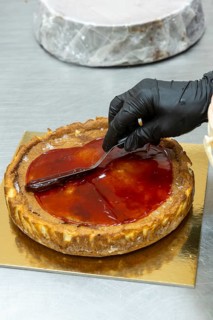 A baker wearing black gloves skillfully spreads glossy red glaze over a freshly baked tart The tart