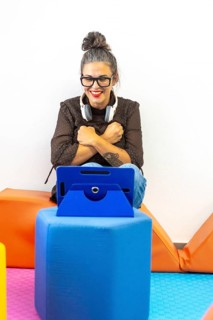 A woman teaching sign language through a video call, sitting on colorful cushions with a tablet