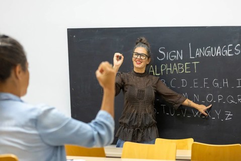 An instructor teaches sign language, pointing to an alphabet chart on a chalkboard A student