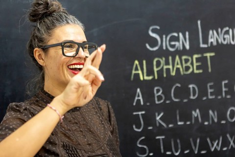 A woman teaches sign language alphabet in a classroom, gesturing with a smile Chalkboard filled