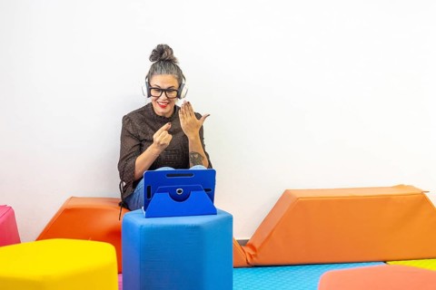 A woman with headphones and an tablet teaches sign language online, demonstrating gestures
