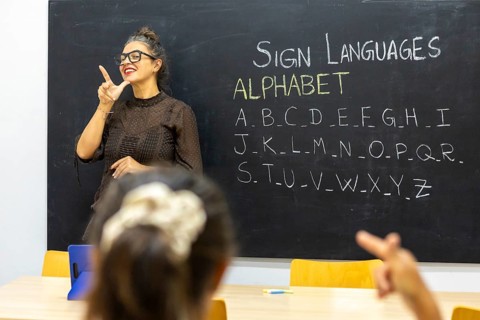 A sign language instructor demonstrates letters from the alphabet on a blackboard in a classroom